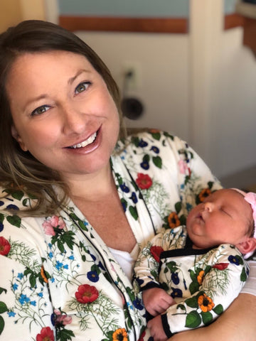 A smiling woman holds a newborn baby. They are are dressed in bamboo pajamas covered with bright garden florals. 