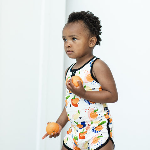 A toddler is holding oranges while wearing a color citrus tank and short set. 