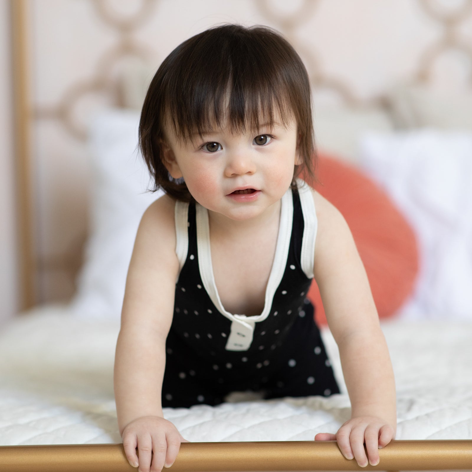A baby. leans against the end of a bed wearing a black tank romper with white polkadots
